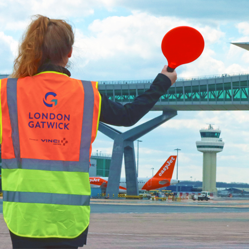 a photo of a ground handler with aircraft guidance paddles, with the pier 6 bridge and control tower in the background